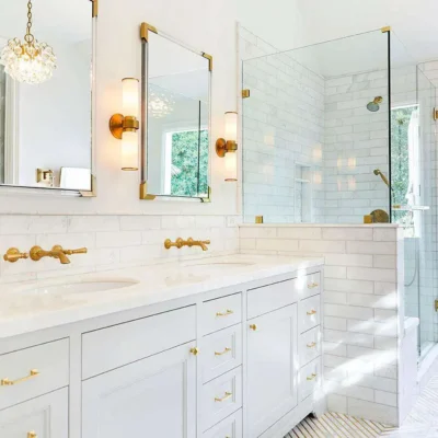 Elegant white bathroom with marble countertops, dual vanities, and gold fixtures. Features a glass-enclosed shower with subway tiles and large windows allowing natural light.