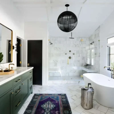 Bright bathroom with a freestanding tub, green cabinetry, gold accents, and a glass-enclosed shower. Features double vanities, black spherical pendant light, and a colorful rug.